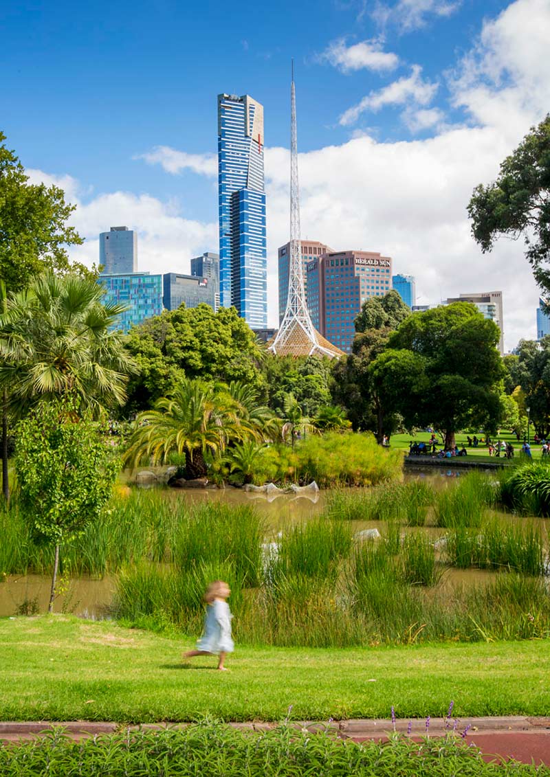 A child runs beside an ornamental pond set among the lawns and trees of the landscaped gardens. The city including the Eureka Tower and Arts Centre are nearby in the background.