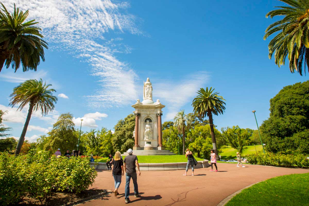 The Queen Victoria memorial statue is positioned at the highest point in the gardens, surrounded by a wide walking path and several palm trees.