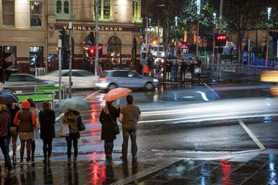 People holding umbrellas on a rainy evening, waiting to cross the road near the Young and Jackson pub.