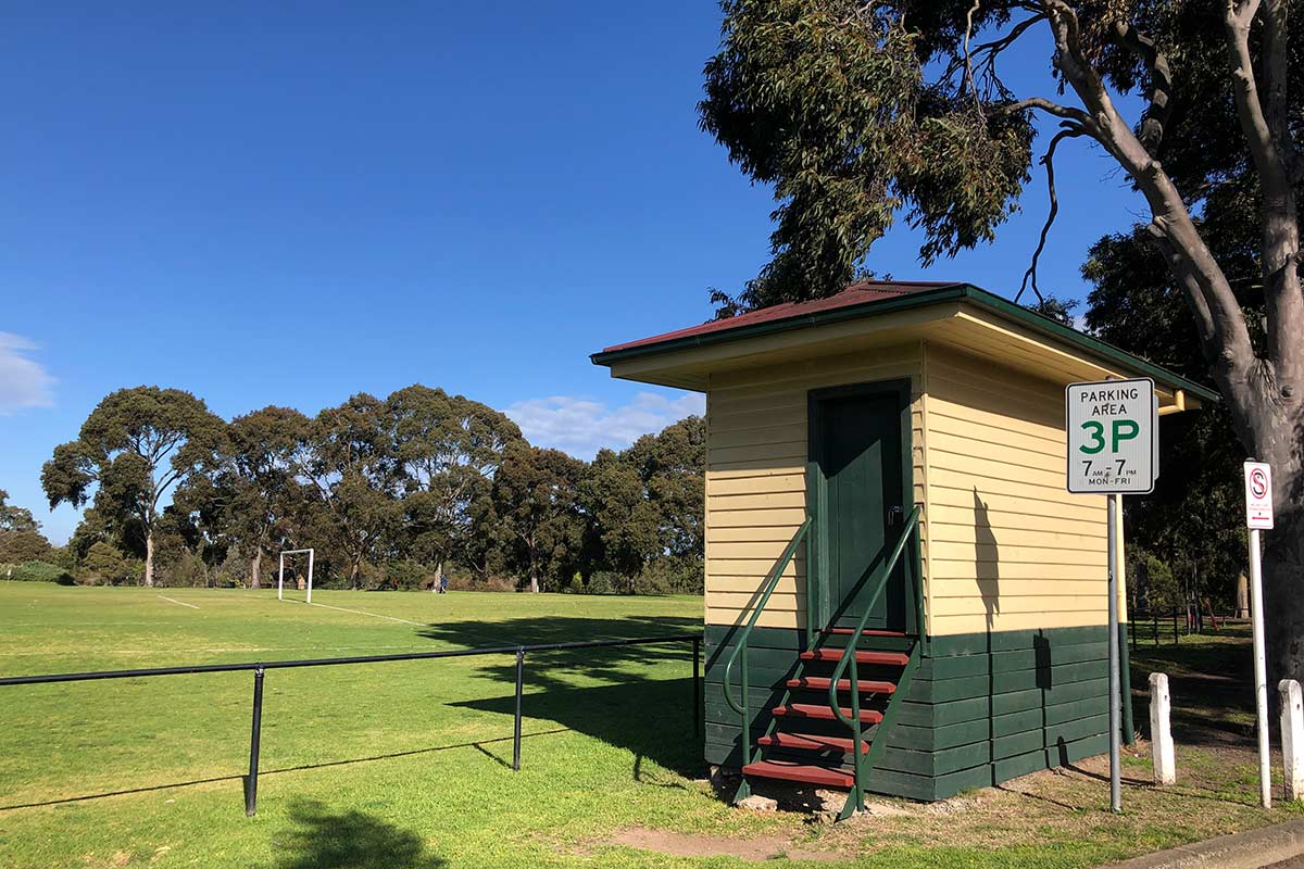 The scorebox, on the edge of the oval and next to the road, is a tiny wooden building with dark green and cream weatherboards, red roof,  and steep steps leading to the door.