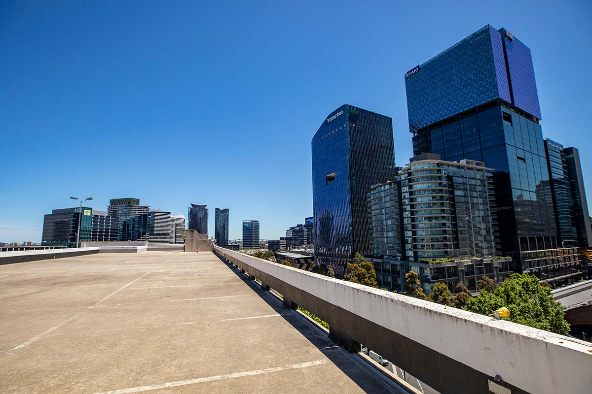 Empty rooftop space with concrete and parking spaces marked in white. Tall city buildings can be seen nearby.