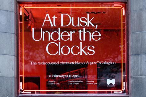 Close-up of red sign with white writing 'At Dusk, Under the Clocks'