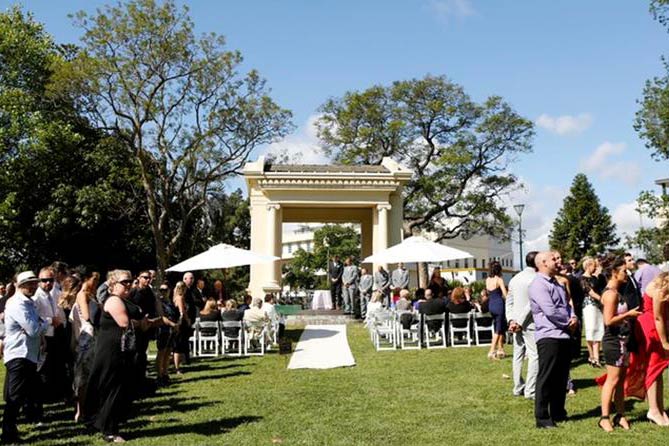 Wedding party and guests standing around a small classical style structure in Fitzroy Gardens.