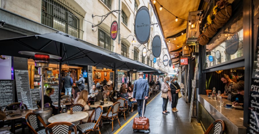People standing and walking in a narrow laneway; cafes and tables line the pavement.