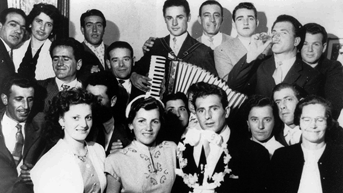 An informal black and white photograph of a wedding party with a cake, accordian player and people drinking.
