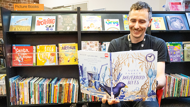 A person sitting in front of a bookshelf with a picture book open in his hands. He is smiling.