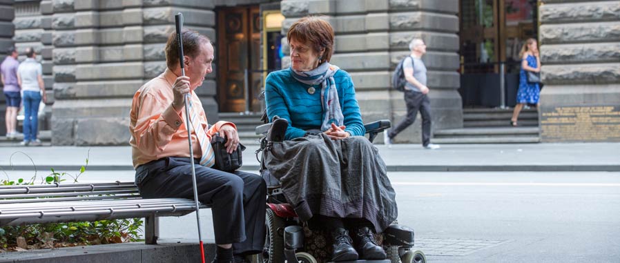 Two people, one with a vision impairment who is seated with a cane and the other in a wheelchair. They are talking to each other in front of the town hall.