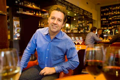 Man in a blue shirt sitting on a lounge chair in front of a bar. Two glasses of red wine and one of white are on a table in the foreground