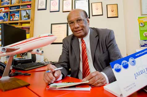 Man sitting at a red table covered with flight tickets, brochures and a model aeroplane. Framed certificates are on the wall behind him.