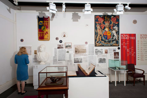 Woman in blue looking at wall of gallery featuring a white bust, brightly coloured tapestries, photos, large open book and glass display cabinet, alongside other ephemera