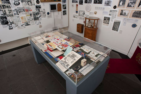 Glass display cabinet showing many brochures and other ephemera next to a white wall covered with photographs and newspaper clippings