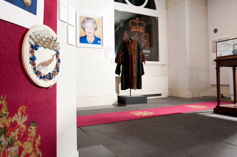Rpyal Melbourne exhibition featuring Lord Mayoral robe with fox fur trim, portrait of Queen Elizabeth, red carpet and embroidery hoop