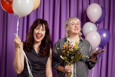 Two people with flowers and baloons in front of a purple curtain