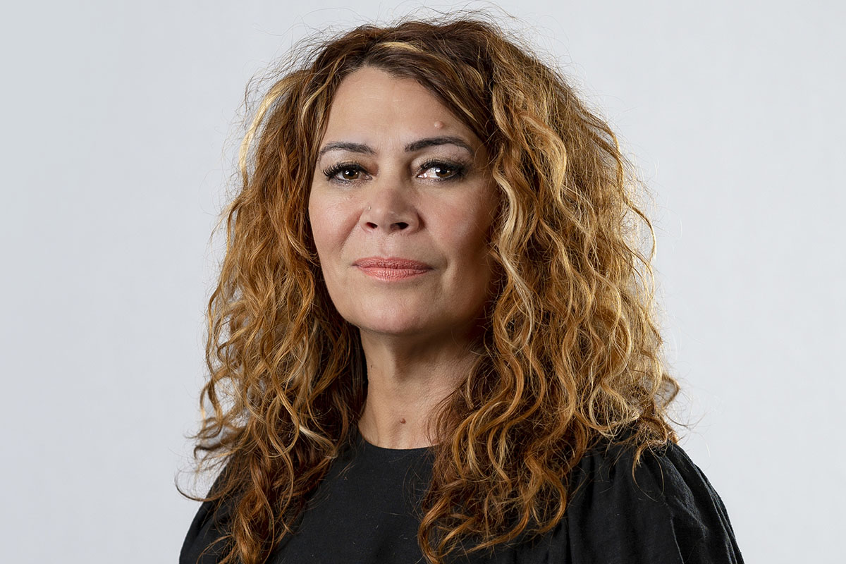 Antoinette Braybrook, a Kuku Yalanji woman, is the keynote speaker at the 2023 National Reconciliation Week Oration. She has curly light brown hair, wears a black top and is standing in front of a light grey background looking towards the camera.