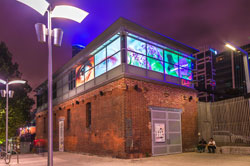 A square brick building with illuminated artwork displayed on its walls. It is night.