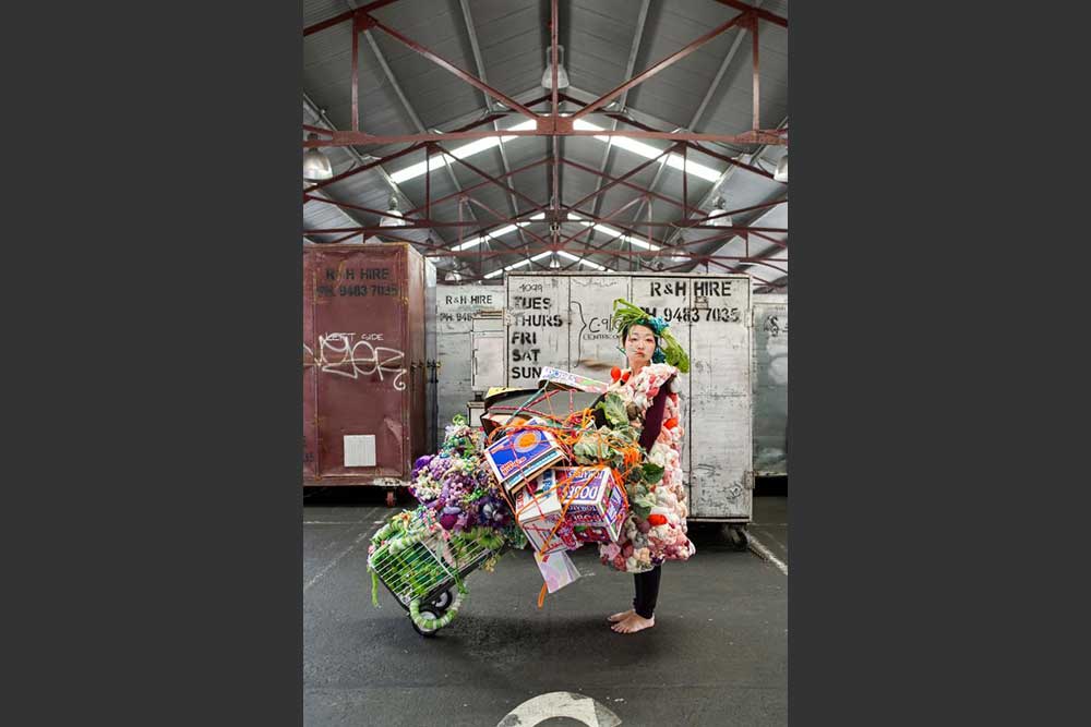 A woman wearing an artwork and holding an overflowing trolley of fruit boxes and fabric while standing in the middle of a Shed at the Queen Victoria Market