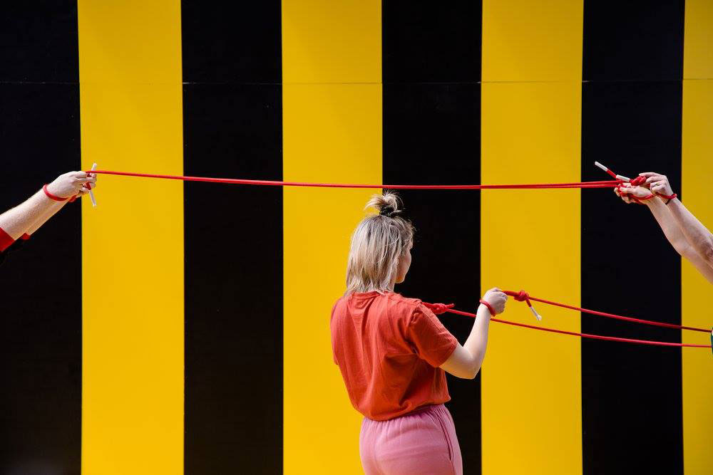 Girl with rope and two pairs of hands holding a straight piece of rope in front of a black and yellow striped wall.