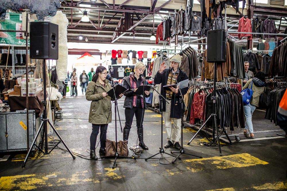 A busy marketplace in the centre of Queen Victoria Market, selling clothes and knick knacks, with three people standing in front of microphones singing and reading sheet music.