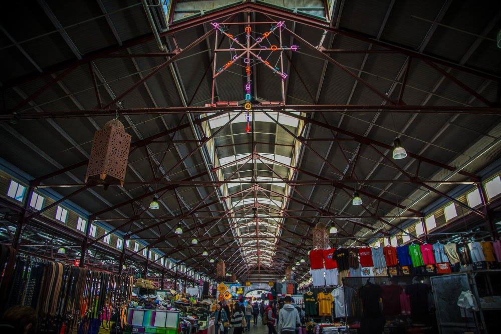 The roof of one of the sheds at the Queen Victoria Market with lots of items for sale in the bottom of the picture. In the top of the roof is a neon artwork in different colours.