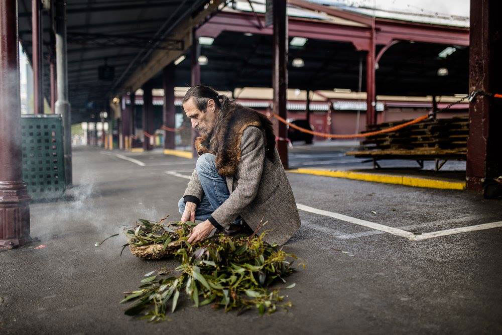 An aboriginal elderon the ground, lighting some eucalyptus leaves for a smoking ceremony, and is wearing a jacket with a possum skin over the top, on the site of the Queen Victoria Market.