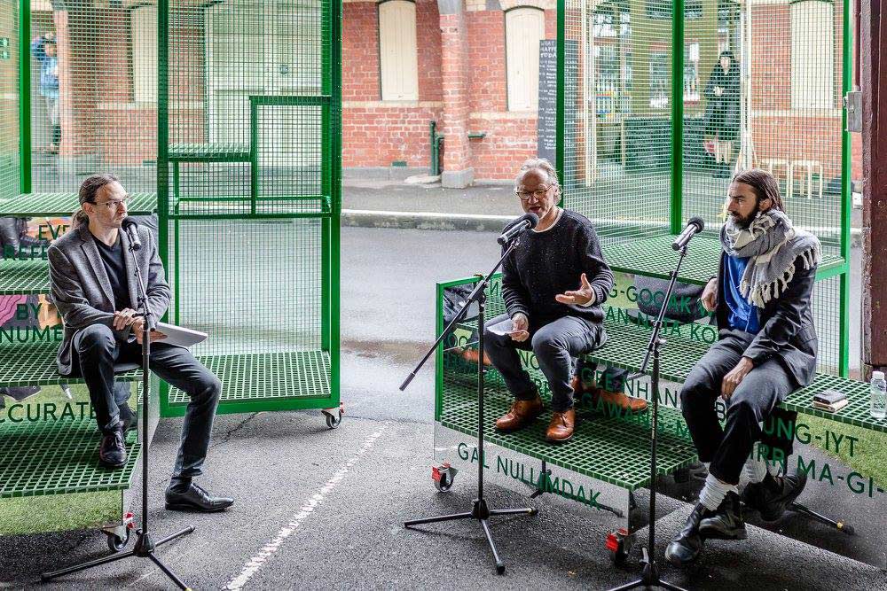 Image of a green cage-like structure opened up with mirrored surfaces that have Woiwurrung words written on them. Three people sit on the sculpture with microphones in front, talking to an audience.