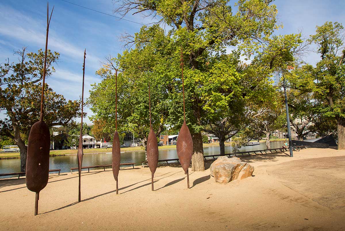 Aboriginal artwork of five metal shields on stakes sticking out of the gravel ground. Mature trees and the Yarra River can be seen in the background.