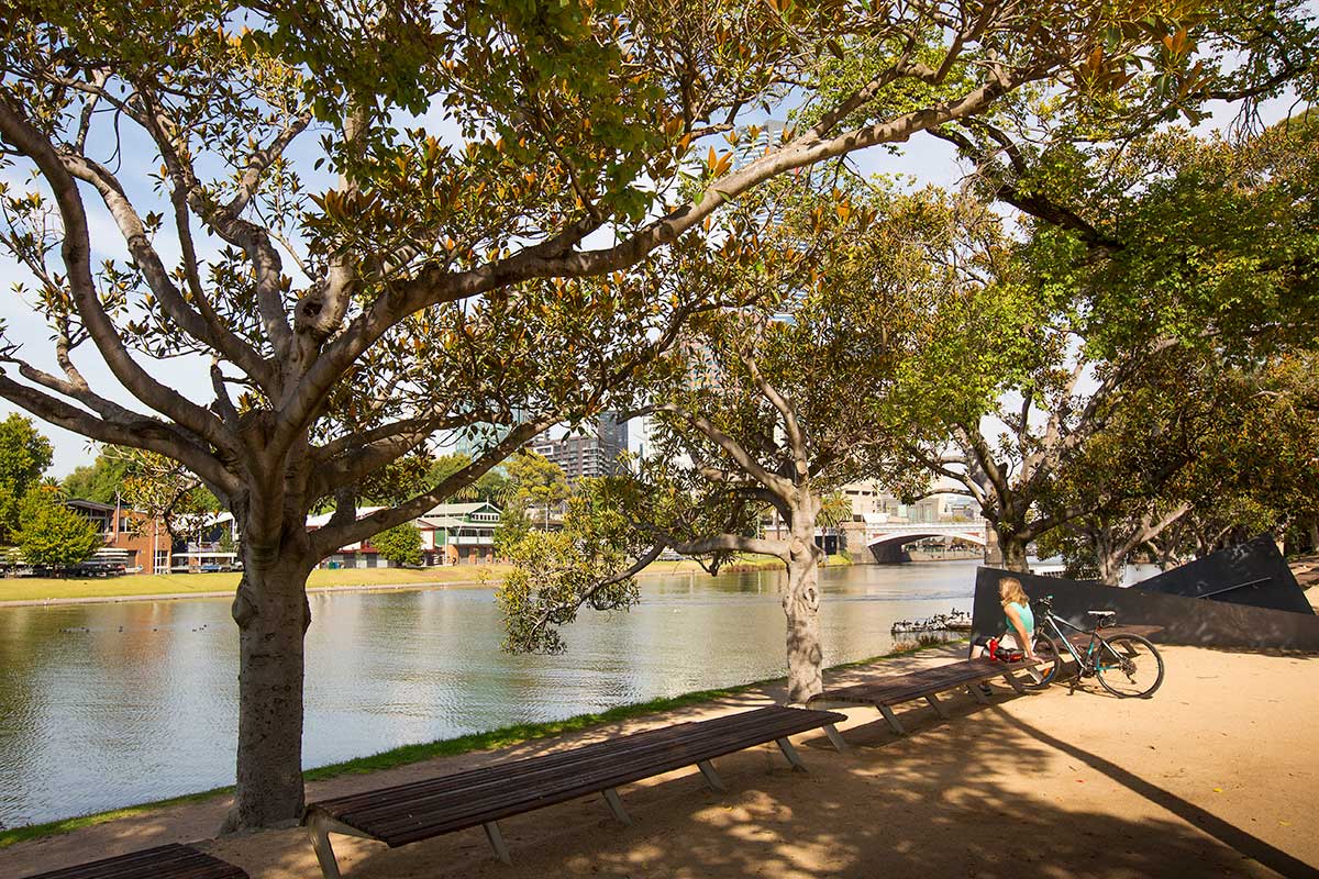 A cyclist rests on a bench under mature trees next to the Yarra River. Boatsheds are visible across the river with Princes Bridge in the background.