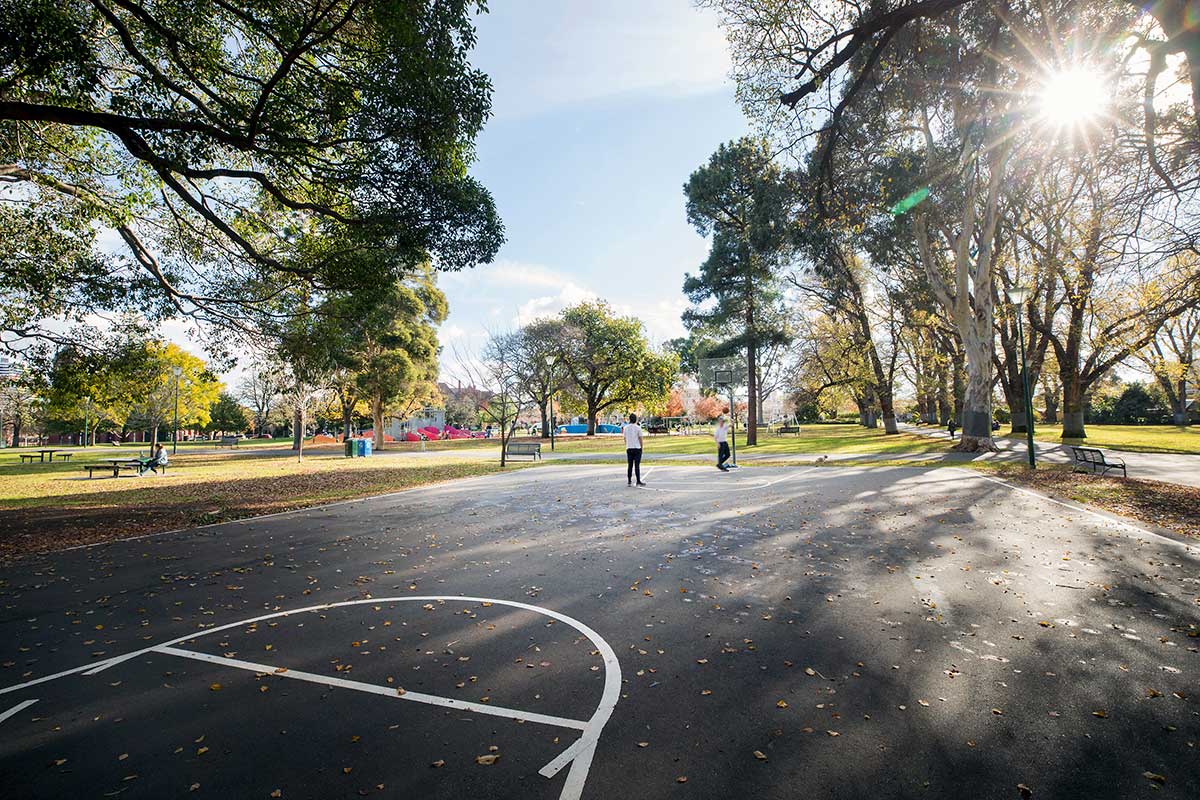 People using the basketball courts in Carlton Gardens. There are many trees surrounding the court, and seats nearby.