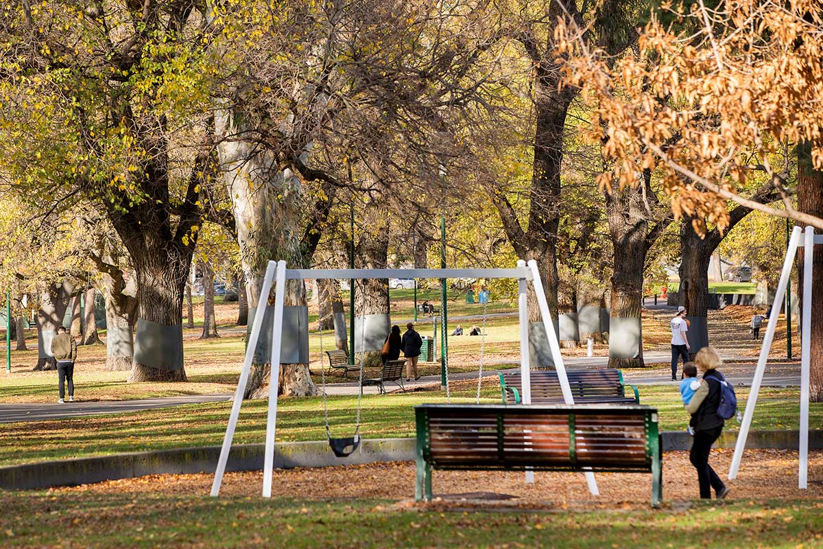 Children's playground in Carlton Gardens with swings, several seats and large elm trees nearby