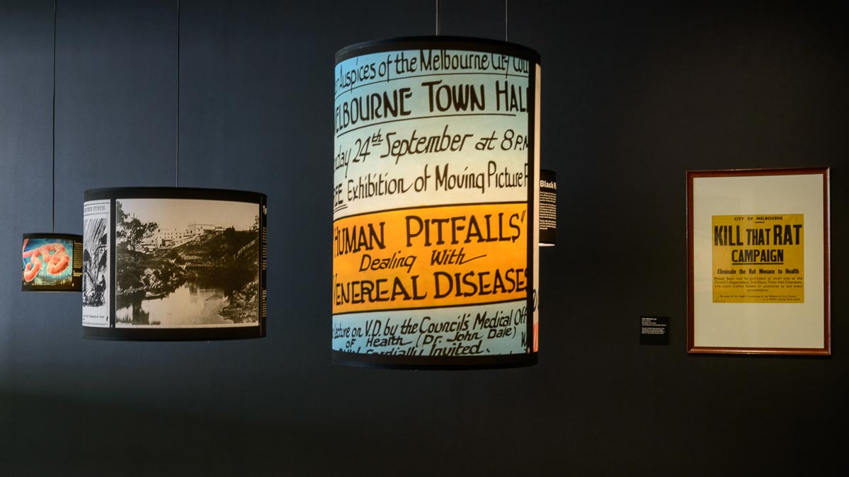 Exhibits on suspended light shades: black and white photo of river and buildings;  signs for 'Human Pitfalls - dealing with venereal disease' exhibition and 'Kill that rat campaign'