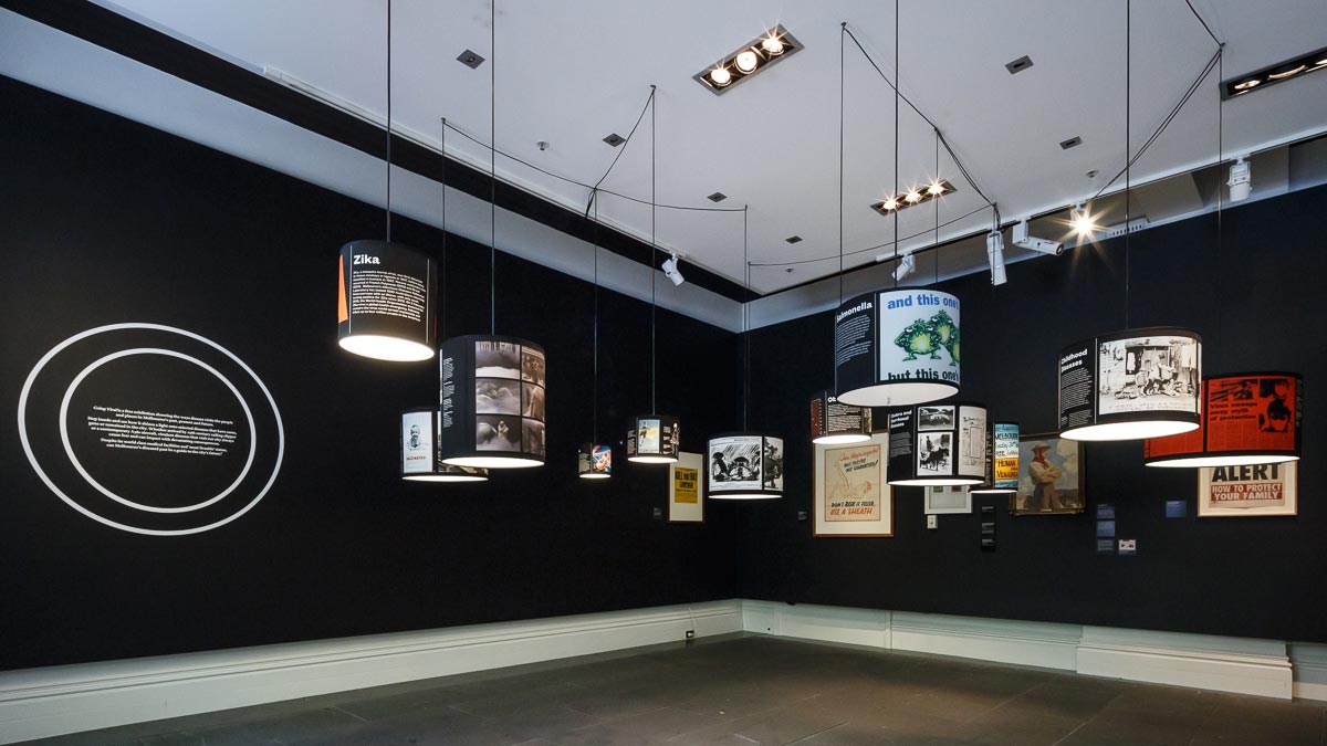 Gallery space with black walls; various exhibits in frames on the wall and presented on pendant lamp shades hanging from the ceiling