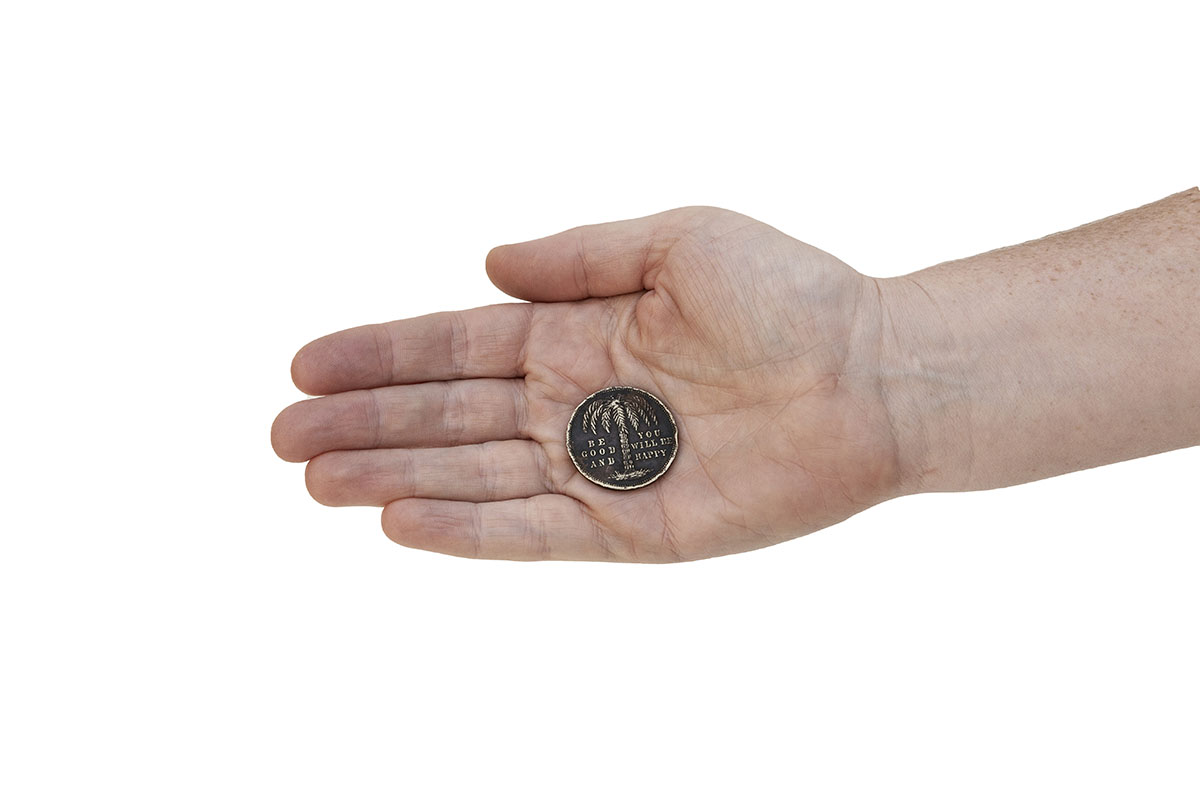 A hand holding a coin that has a slogan and a palm tree stamped on it. The slogan reads ‘BE GOOD AND YOU WILL BE HAPPY’.