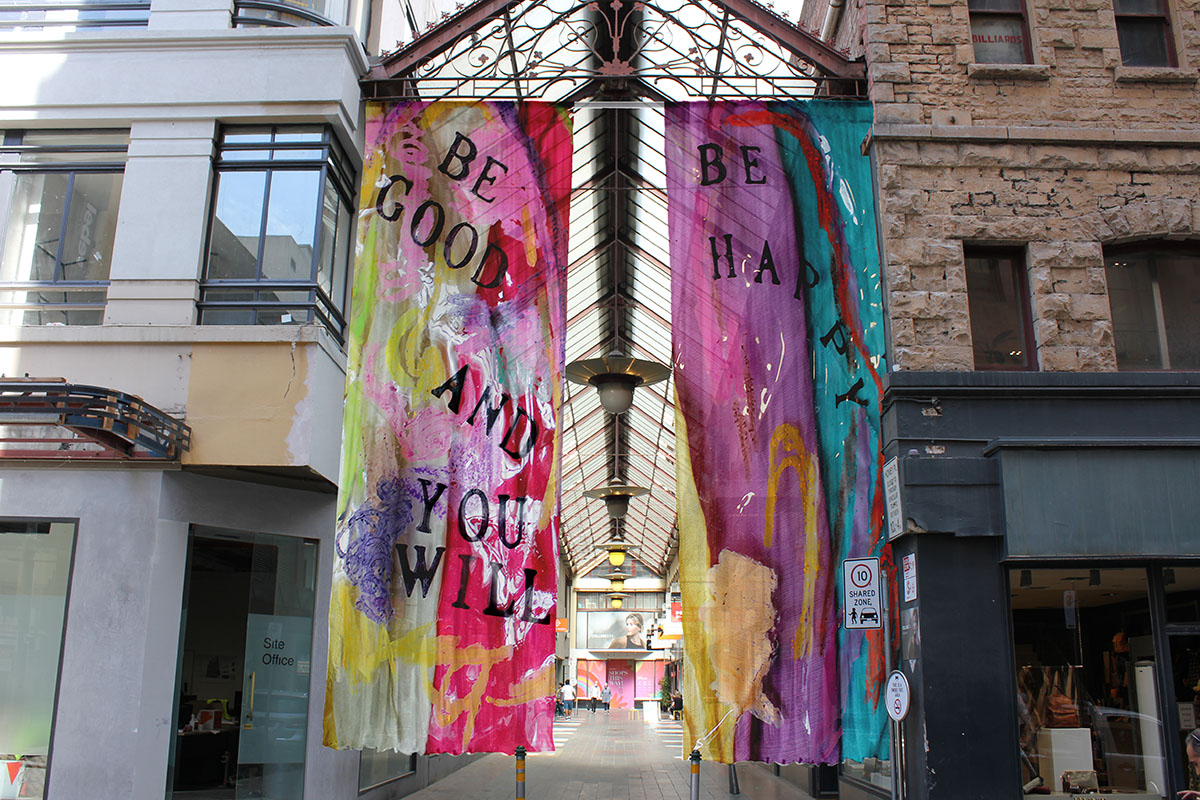 Colourful large curtains hang from a steel structure covering Howey Place laneway with the words ‘BE GOOD AND YOU WLL BE HAPPY’