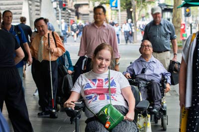 Two people in wheelchairs and a person with a cane among a crowd of people in the city