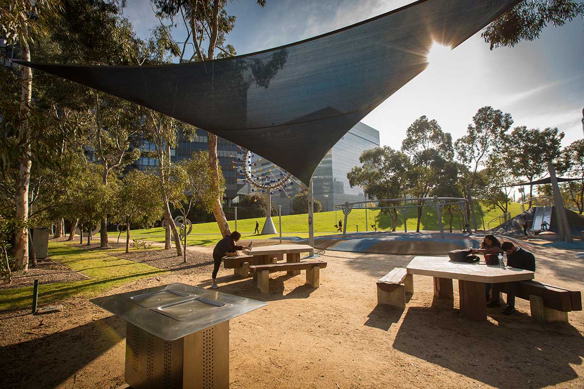 Picnic tables and seats under a shadecloth, near the playground and a sculpture.