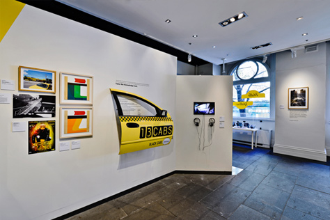 Close-up of yellow taxi door mounted on the gallery wall plus five small paintings/photos