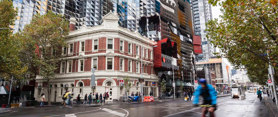 City street scene with pedestrians and cyclists in front of an older heritage building, with a modern RMIT building behind it