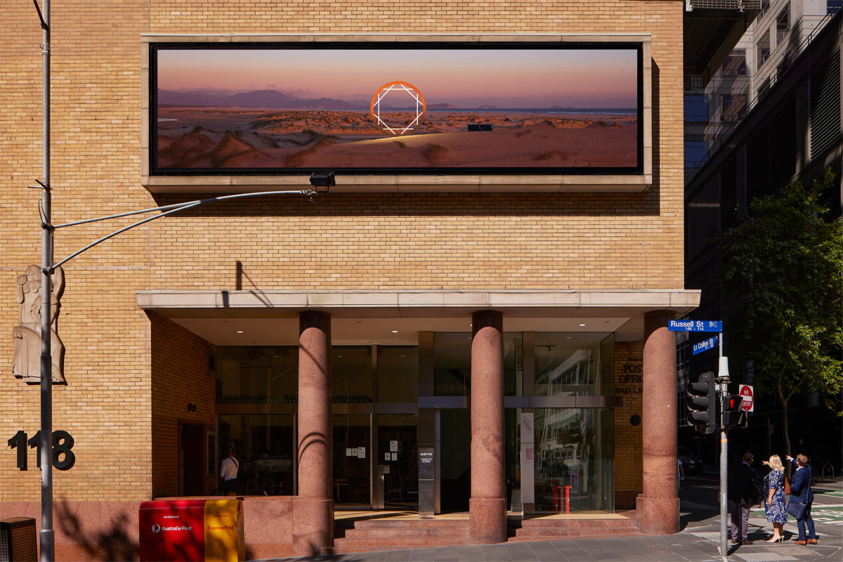 Closer view of the artwork which comprises a panoramic photo of a desert landscape with pink sunset sky. A representation of a camera aperture overlaps with the horizon in the centre of the photo.