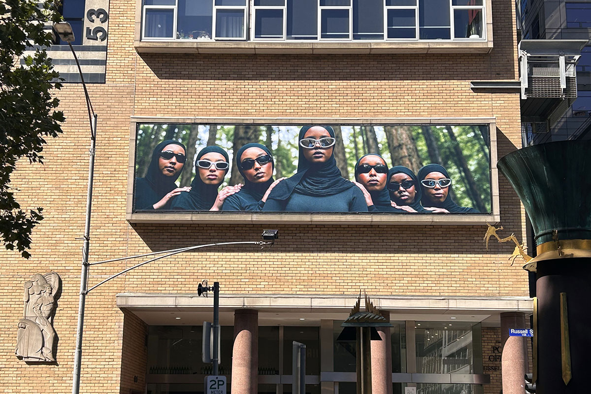A large billboard image on an orange-brick building depicting a group of women wearing hijabs and sunglasses.