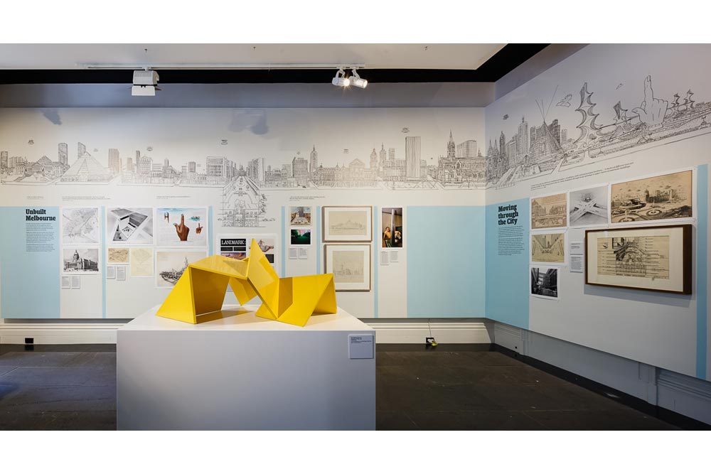 Scale model of the yellow 'Vault' sculpture, other exhibits displayed on walls in the exhibition space and black line drawing/mural of existing and imaginary buildings of Melbourne's cityscape