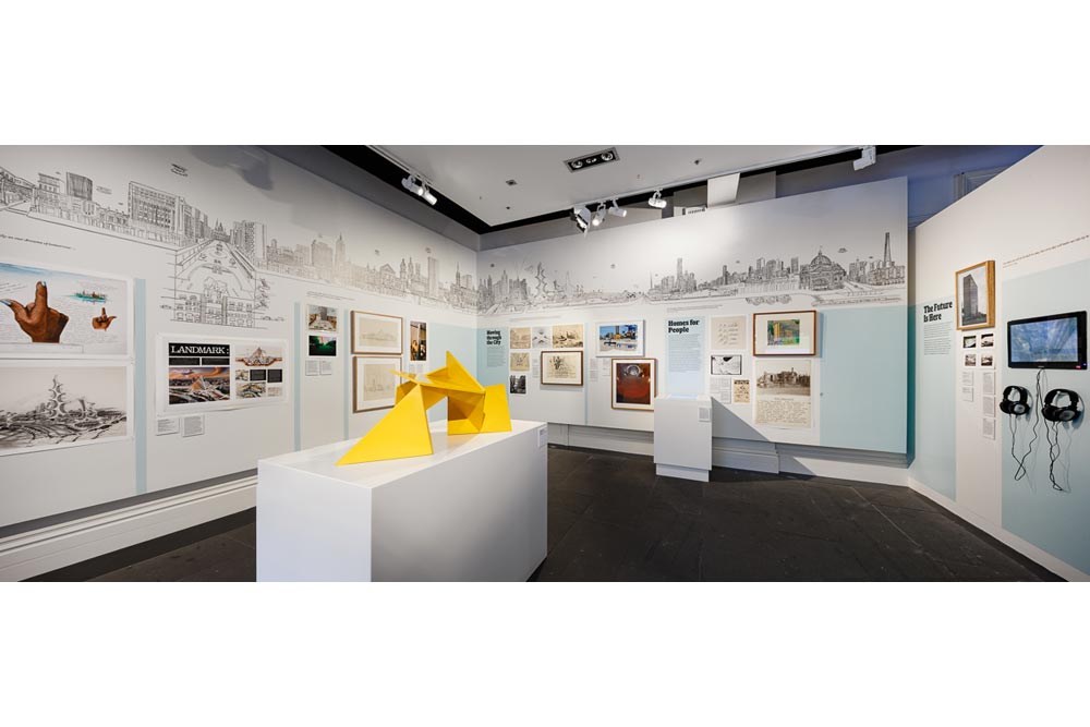 Exhibition space showing the yellow 'Vault' sculpture scale model in the centre and plans, posters, maps, mural and other exhibits on the surrounding walls