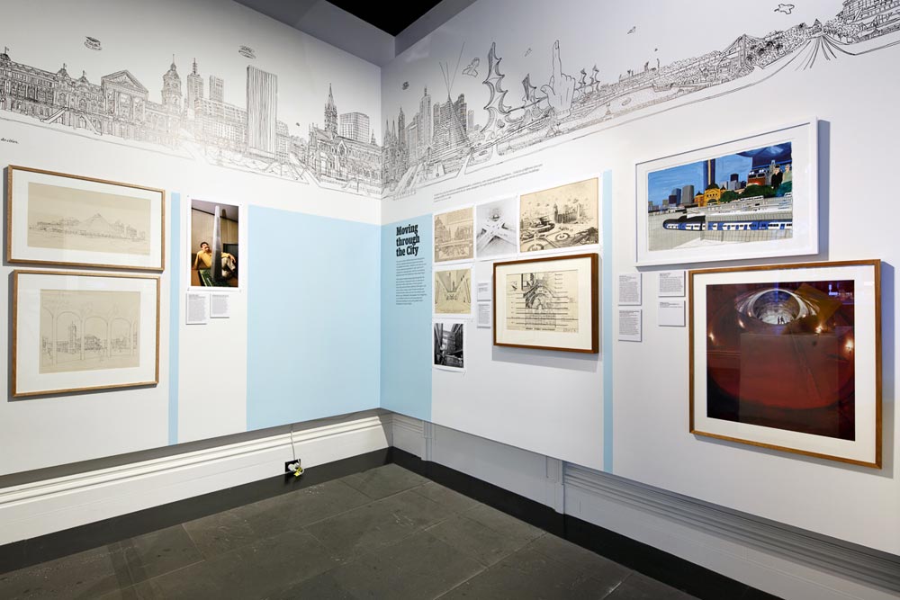 View of one corner of the gallery space with various pictures, photos and plans exhibited underneath a section of the mural