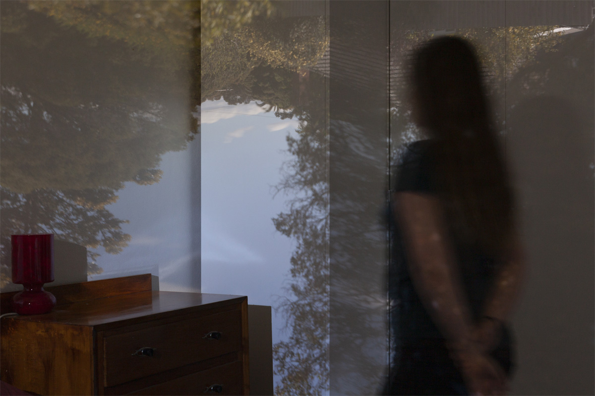 A person standing inside a bedroom looking at an upside down photograph of a garden projected onto the wall.