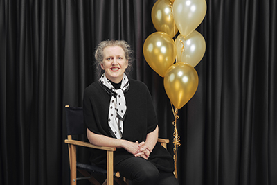 A person seated in front of a velvet curtain and balloons. 