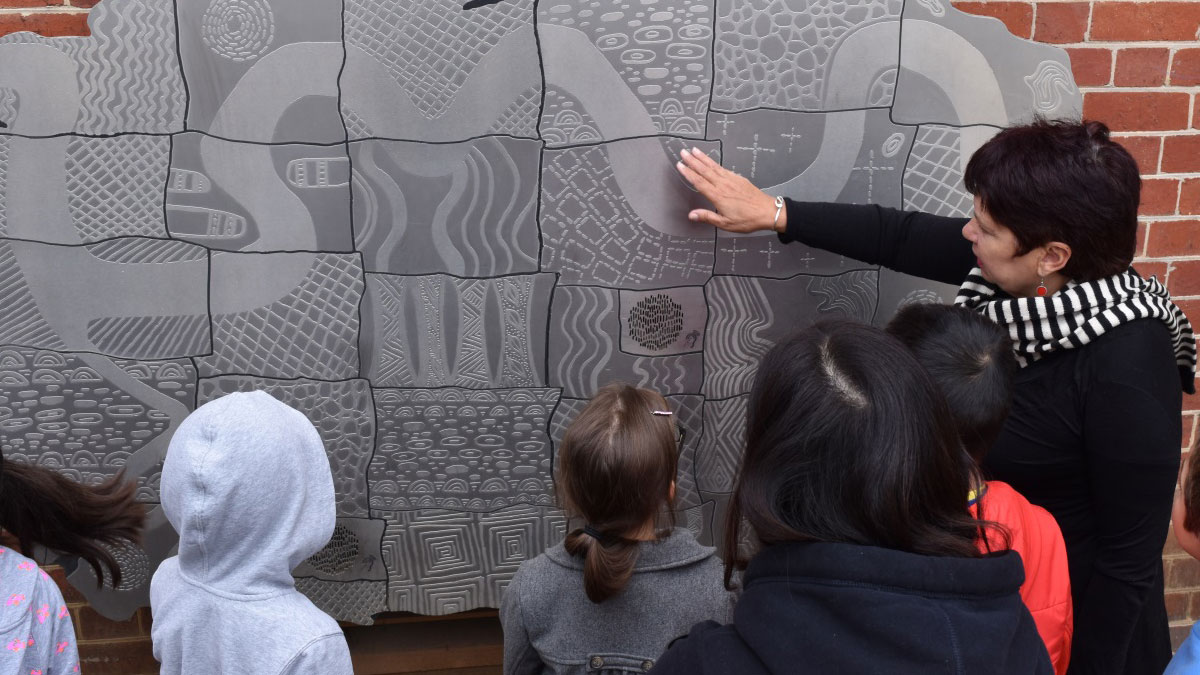 A woman stands in front of a group of children and gestures to an artwork on a wall.