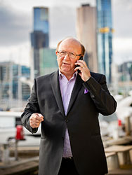 Ahmet Ali talking on the phone outdoors with city buildings and waterfront in the background.