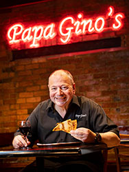 Alex Brosca in his restaurant, holding a slice of pizza and glass of wine and standing under a red neon 'Papa Gino's' sign.