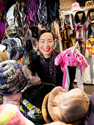 Iris Chan Zhang standing among hats and scarves in her market stall, holding a novelty pink unicorn hat.