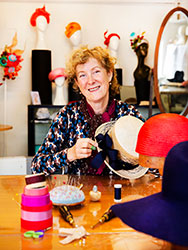 Louise Macdonald in her studio with millinery equipment and colourful hats displayed on mannequins.