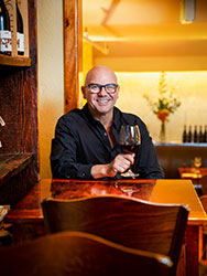 Michael Tenace sitting at a small wooden table in his restaurant with a glass of red wine.